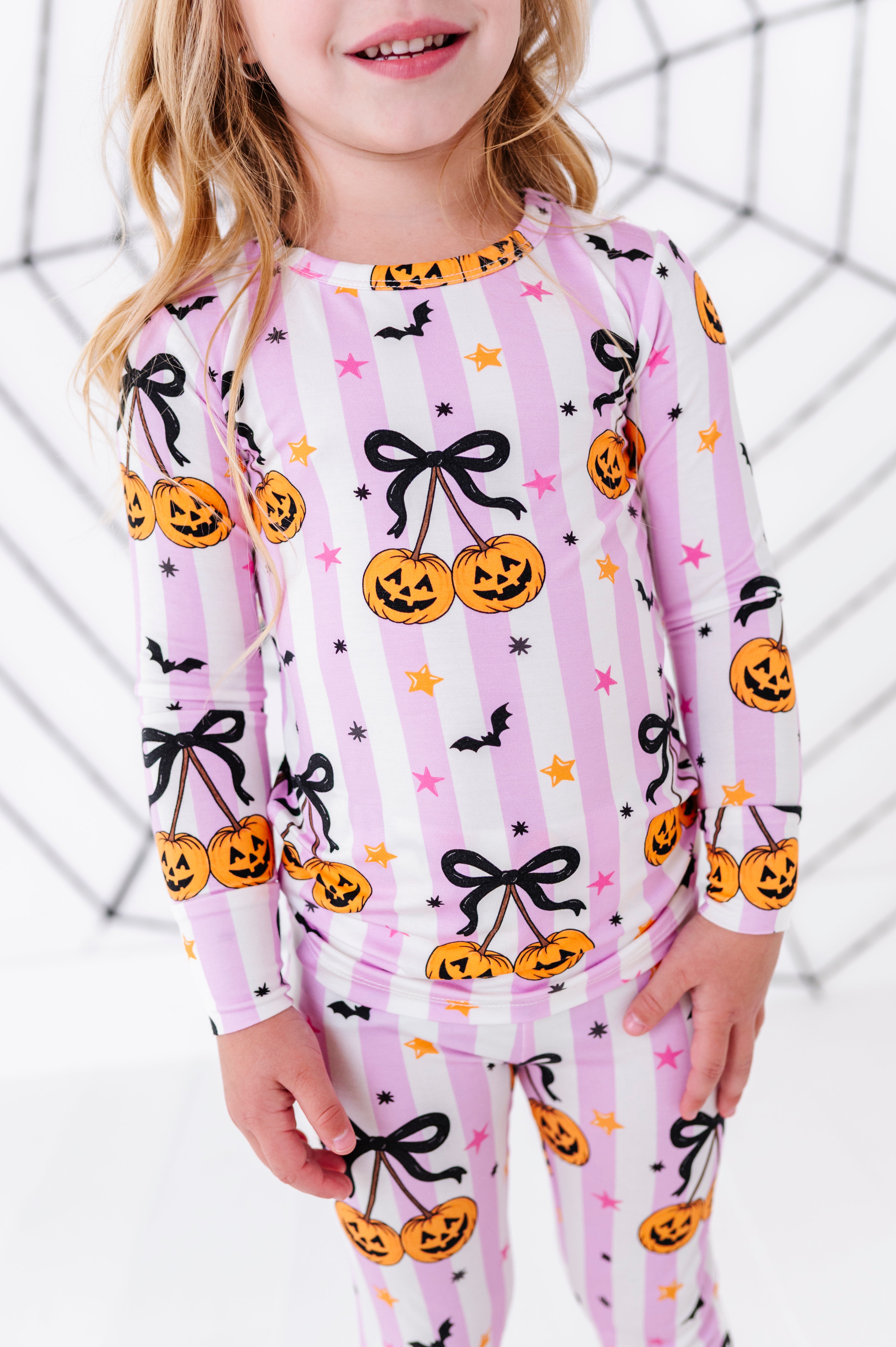 A young girl wearing a pink and white striped long-sleeve pajama top and matching leggings with a Halloween print of jack-o'-lanterns, black bows, bats, and stars.